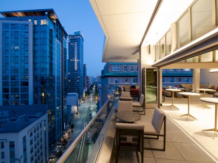 The image shows a modern outdoor terrace with seating, overlooking a cityscape with tall buildings during dusk or early evening.
