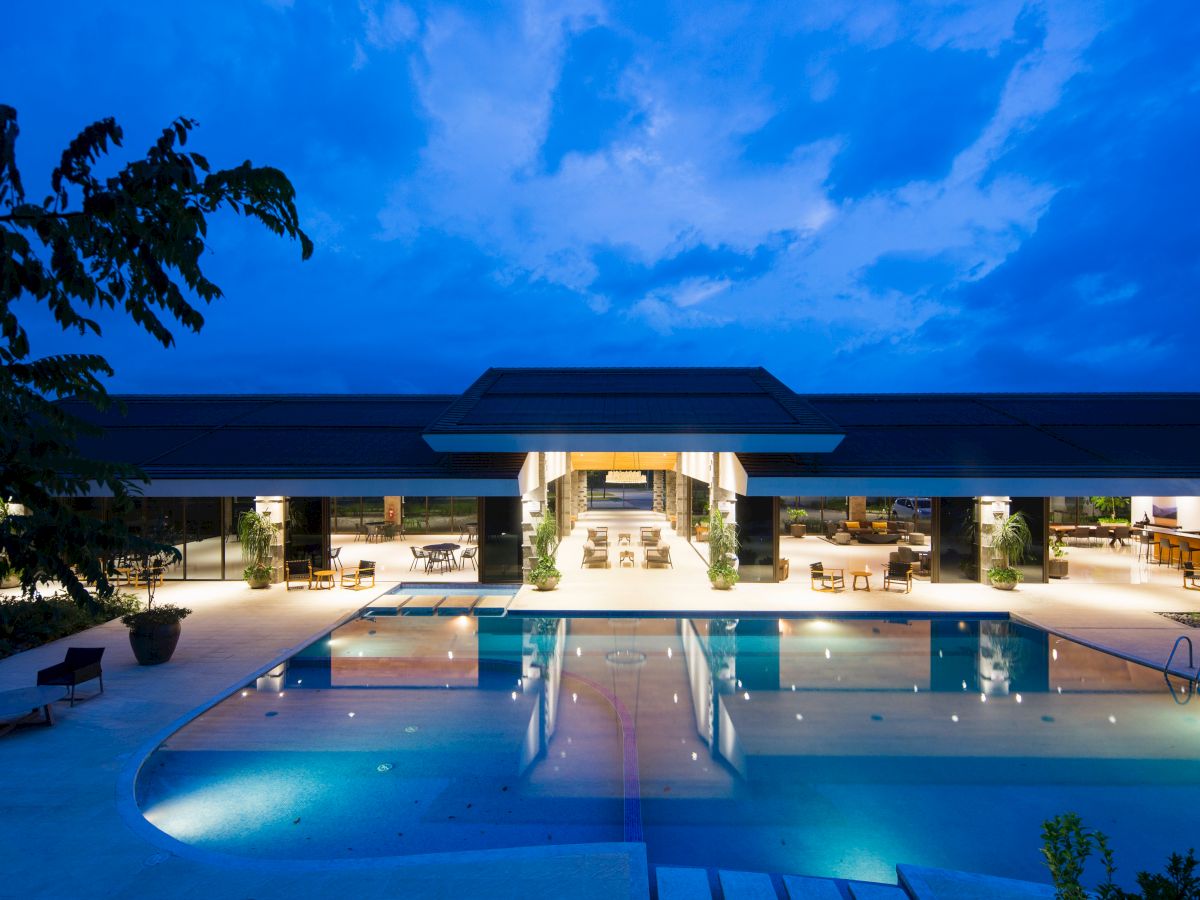 A large, illuminated building with an outdoor swimming pool at dusk, featuring open indoor spaces and outdoor seating areas.