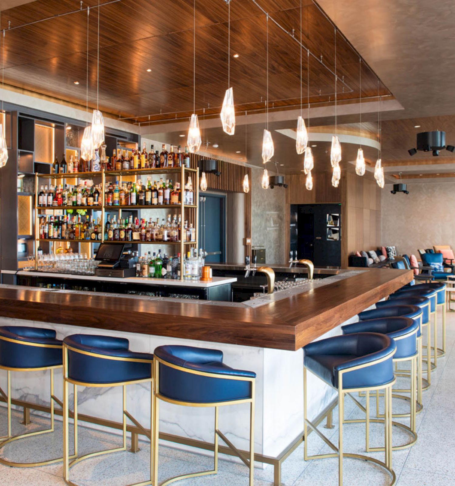 The image depicts a stylish bar with modern decor, featuring hanging lights, bar stools, a well-stocked bar, and a lounge area in the background.