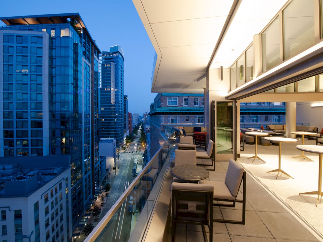 A modern high-rise cityscape is shown, with a view from a balcony. The space includes outdoor seating and tables, with buildings and streets visible below.