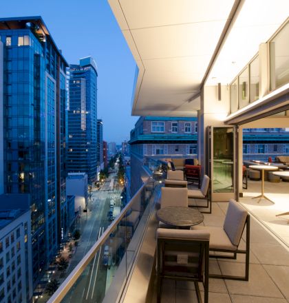 A modern cityscape view from a high-rise balcony at dusk, featuring tables and chairs with tall buildings in the background.