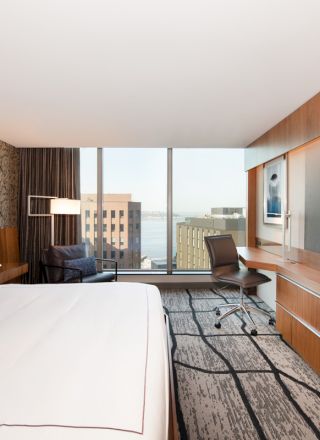 A modern hotel room features a large bed, a desk with a chair, a flat-screen TV, a lounge chair, and floor-to-ceiling windows with a city view.