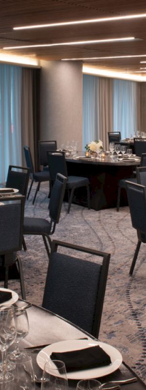 A modern, elegant banquet hall with round tables set with black napkins, glassware, and floral centerpieces, under warm ceiling lights.