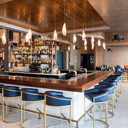 A modern bar with blue seating, hanging lights, and a fully stocked bar counter; cozy seating areas and tables are visible in the background.
