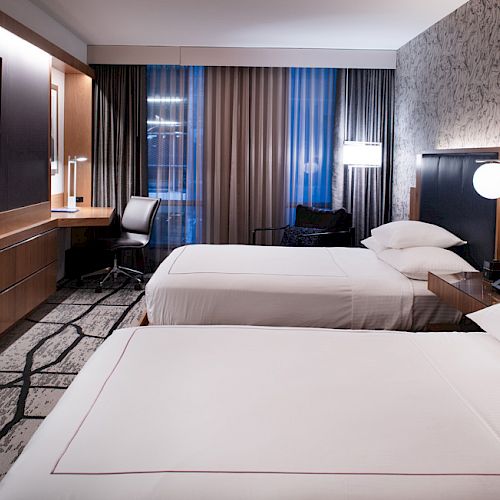 The image shows a modern hotel room with two neatly made beds, a flat-screen TV, a work desk with a chair, and ambient lighting.