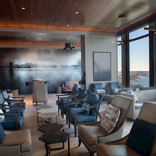 A modern lounge with comfortable seating, large windows offering an ocean view, and contemporary decor including a cityscape mural on one wall.