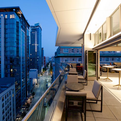 A modern high-rise balcony at dusk overlooking a city street, furnished with tables and chairs, and illuminated by soft lighting.