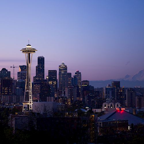 A cityscape at dusk featuring the Space Needle, tall buildings, and Mount Rainier in the background. The sky is a gradient of purple and pink.