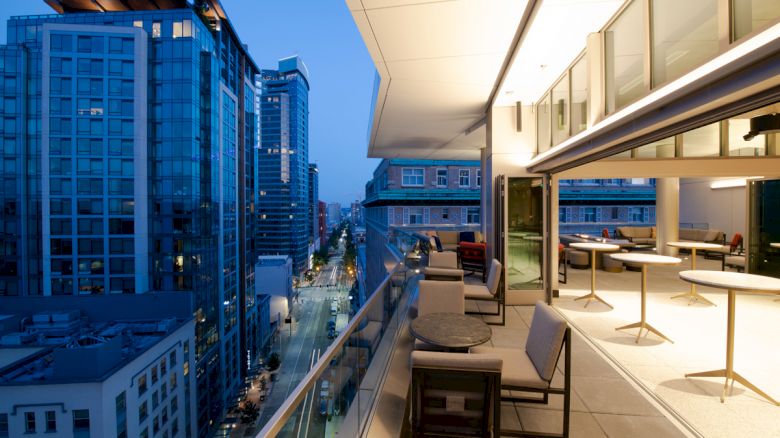 A modern balcony with seating overlooks a cityscape at dusk. The illuminated buildings and evening sky create a serene and vibrant atmosphere.