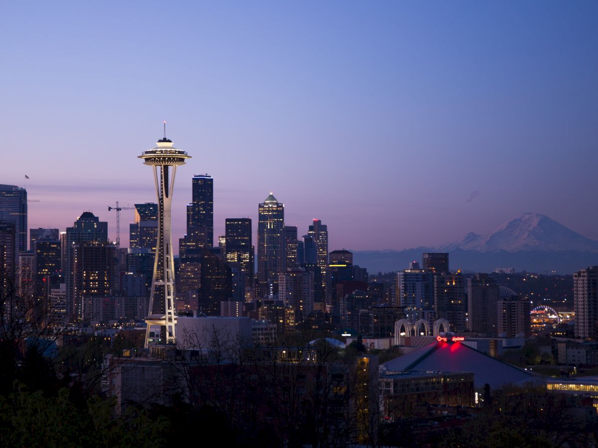 The image shows the Seattle skyline at dusk, featuring the iconic Space Needle and Mount Rainier in the background, with city buildings illuminated.
