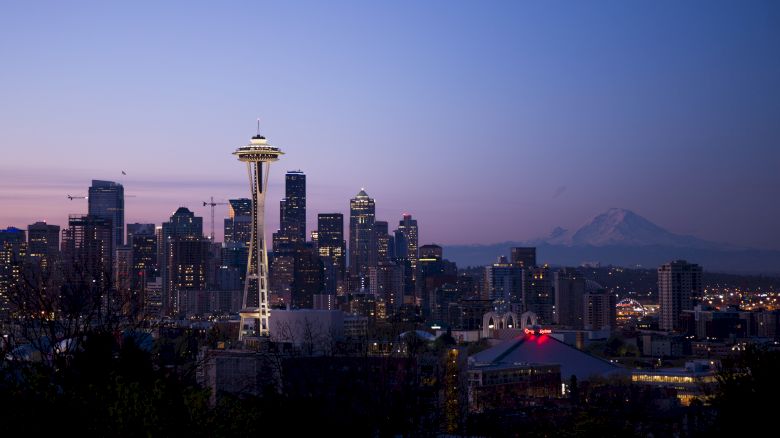 The image shows the Seattle skyline at dusk, featuring the iconic Space Needle and Mount Rainier in the background, with city buildings illuminated.