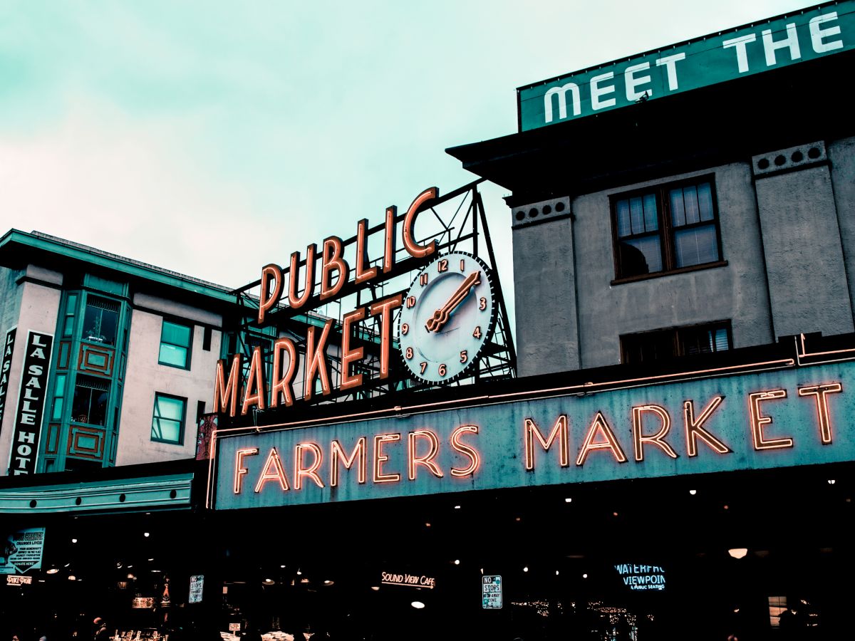 The image shows a public market with neon signs for "Public Market" and "Farmers Market" on the front. An analog clock is also visible.