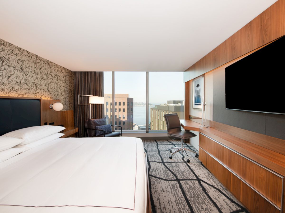 A modern hotel room features a large bed, wall-mounted TV, desk with chair, and a window with a cityscape view.