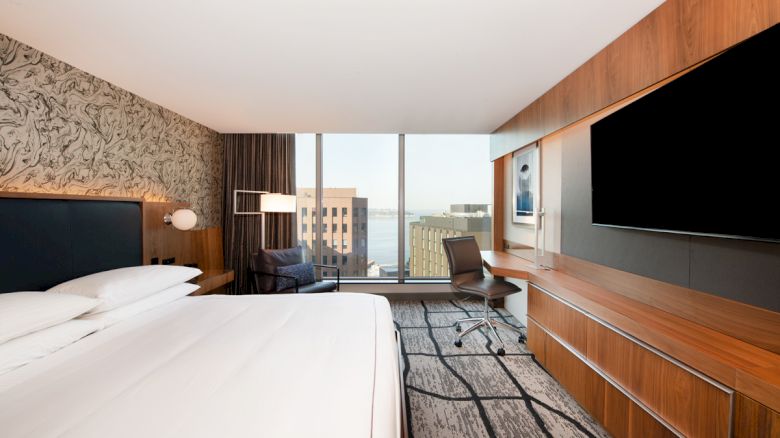 A modern hotel room features a large bed, wall-mounted TV, desk with chair, and a window with a cityscape view.