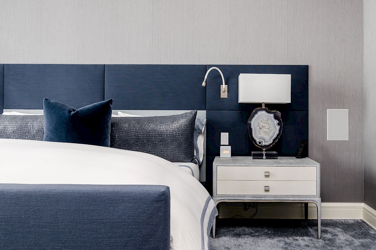 A neatly made bed with blue and white bedding, a navy padded headboard, and a white nightstand with a lamp and decor.