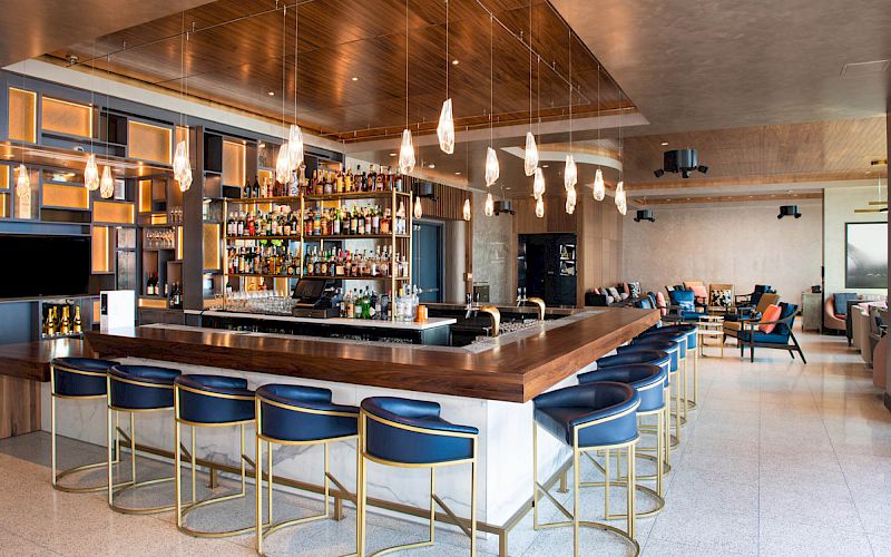 This image shows a modern bar with a well-stocked liquor shelf, blue bar stools with gold frames, pendant lights, and a seating area in the background.