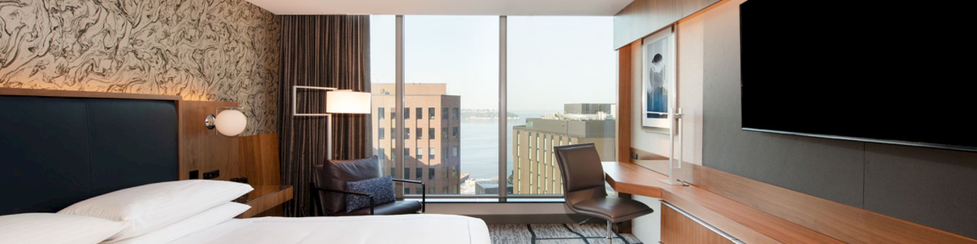 A modern hotel room with a king-size bed, wall-mounted TV, desk with a chair, large window with a city and water view, and contemporary decor.